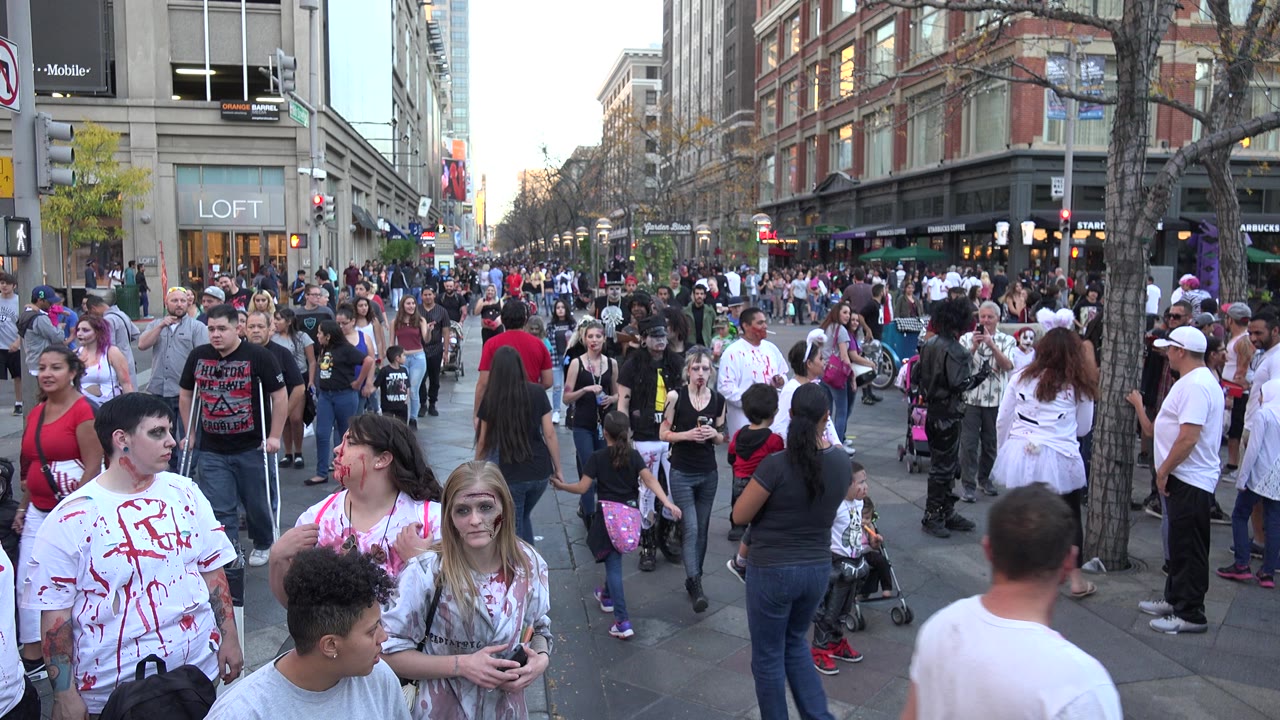 A Denver, Colorado city street is packed with pedestrians, including several costumed people near the foreground and storefronts along both sides of the block.
