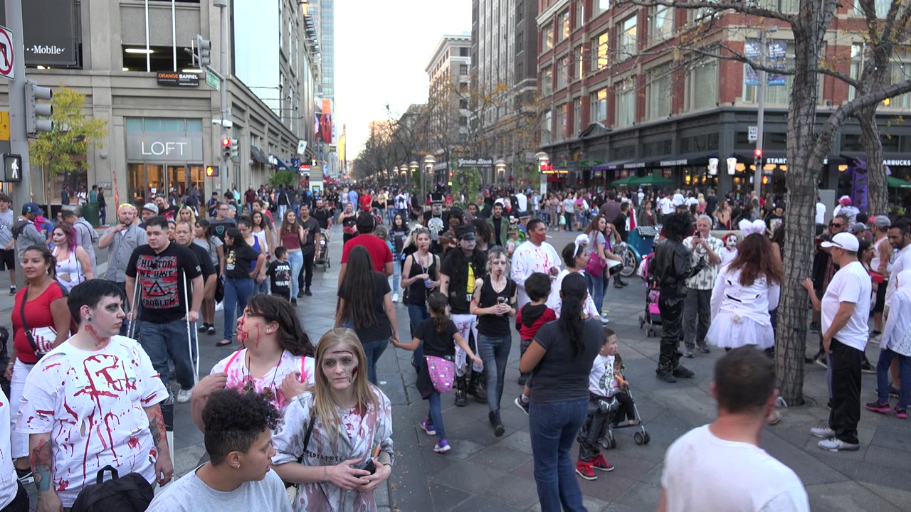Pedestrians continue filling the Denver street, with a person in a white costume near the right side, blood-stained costume clothing in front, and shop windows in the background.