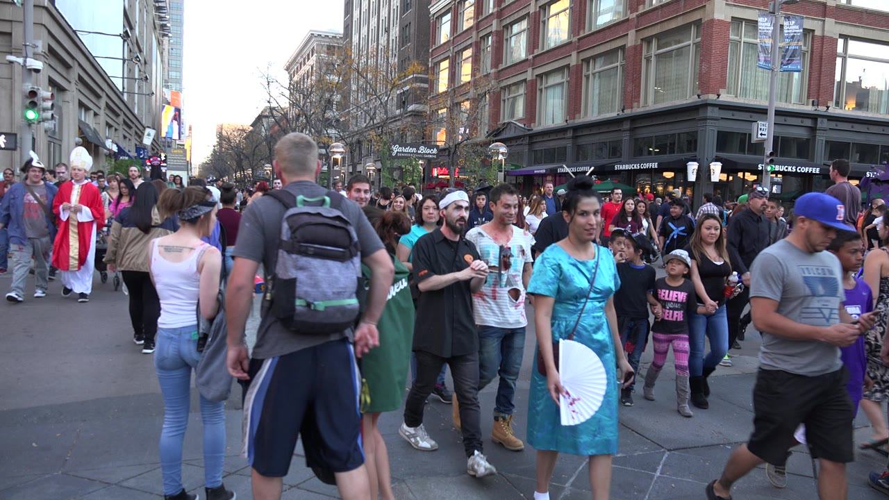Denver, Colorado street crowd with pedestrians walking toward camera, a red-and-white costumed figure at left, storefronts, traffic lights, and visible faces.