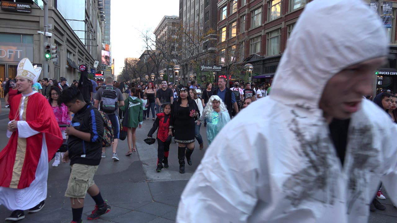 Denver pedestrians move through a downtown street while a red-robed costumed person stands at left and a white hooded person remains close in the foreground.