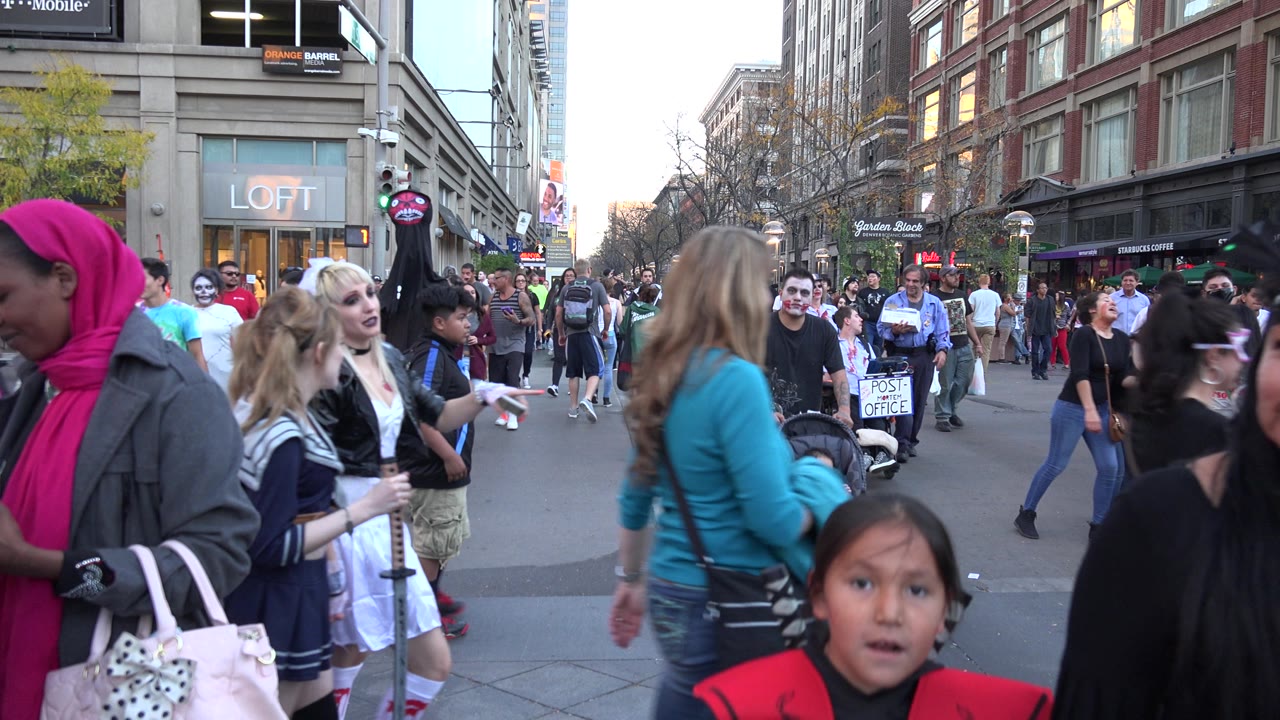 Denver downtown sidewalk scene with many pedestrians, a child near the foreground, storefront signs, traffic lights, and tall commercial buildings.
