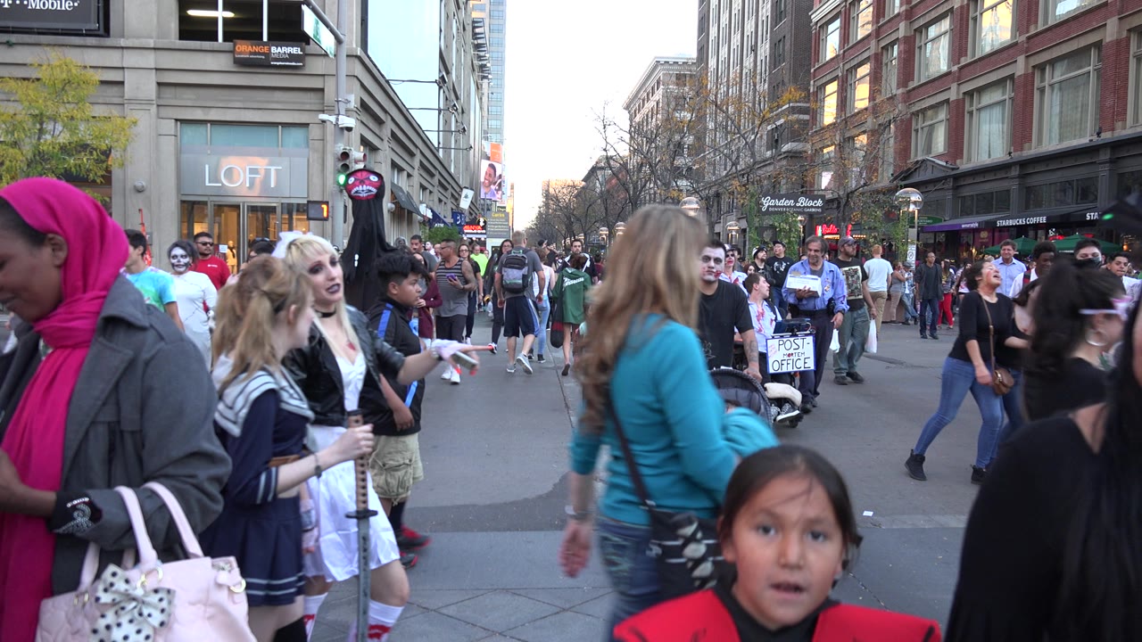 A dense Denver street crowd moves through the frame, with close foreground pedestrians, a child facing camera, and commercial storefronts in the background.