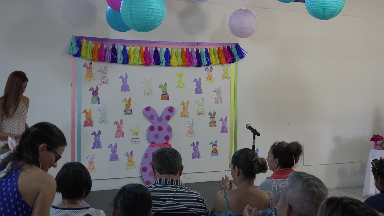 At 0:01 in Cabo San Lucas, Mexico, a woman stands beside the Easter display while seated adults watch the presentation area and microphone stand.