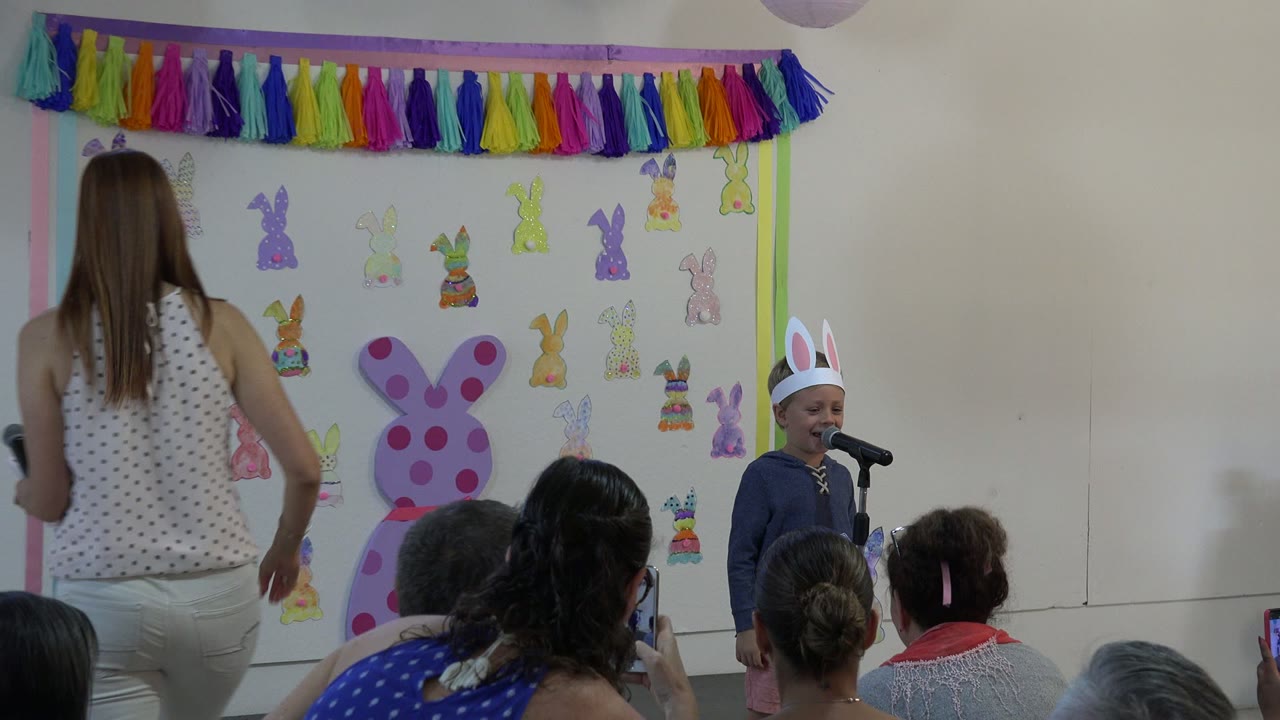 The young boy in paper bunny ears stands at the microphone in Cabo San Lucas while a woman moves away and adults in the foreground record the school performance.