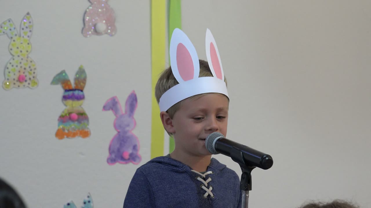 Young child wearing paper bunny ears speaks into a microphone.