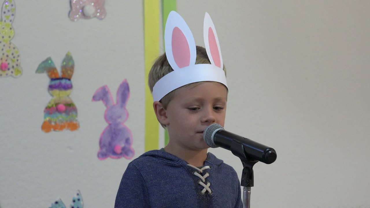 At 0:01, the child looks slightly downward while standing close to the microphone, with a colorful bunny craft and a purple bunny drawing visible behind him in Cabo San Lucas.