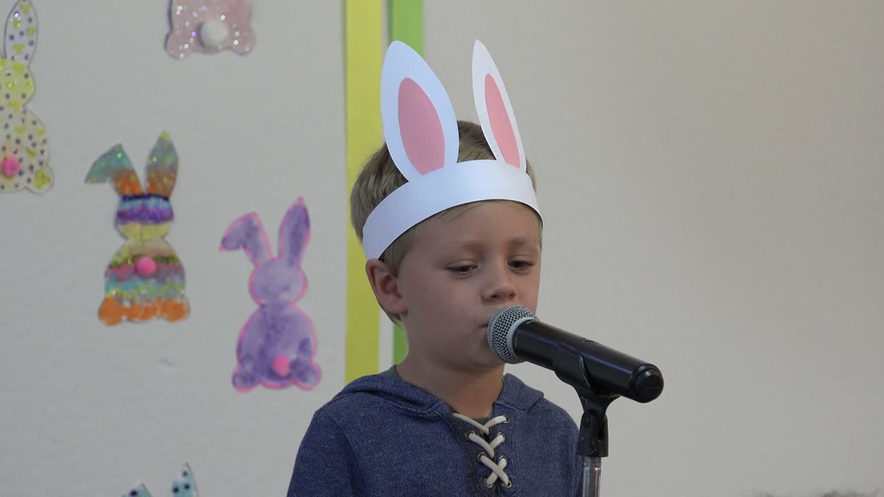 Boy in white rabbit ears speaking into a black microphone indoors.