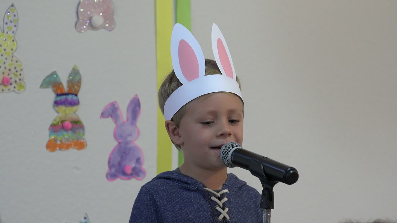 Boy in bunny ears looks downward while standing at a microphone.