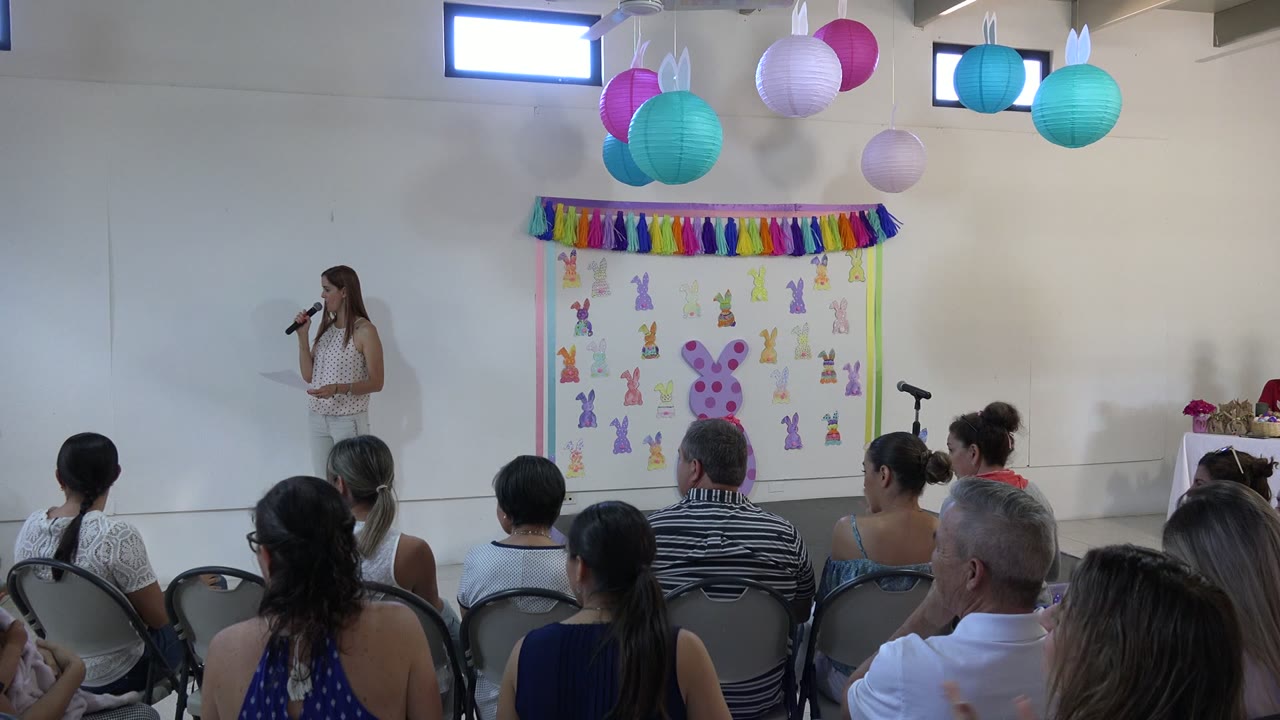 Teacher speaks into a microphone before seated parents in a decorated school room.