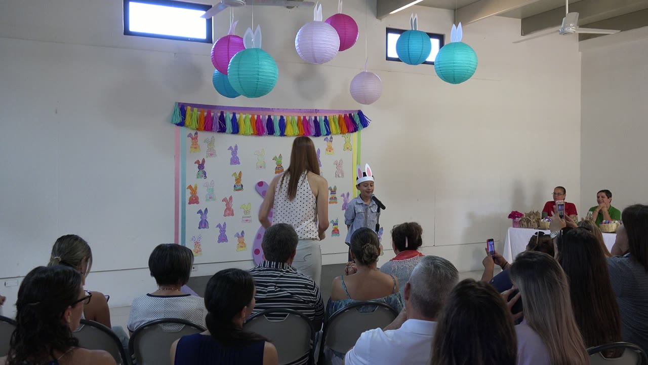 The child remains near the microphone in the decorated indoor presentation area as a woman stands nearby and parents watch from rows of chairs in Cabo San Lucas, Mexico.