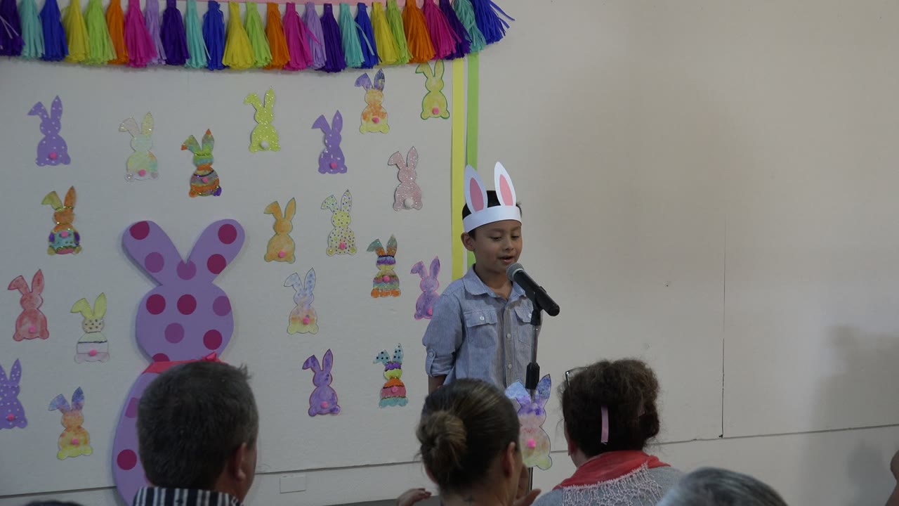 Young boy in bunny ears stands at a microphone before Easter decorations.