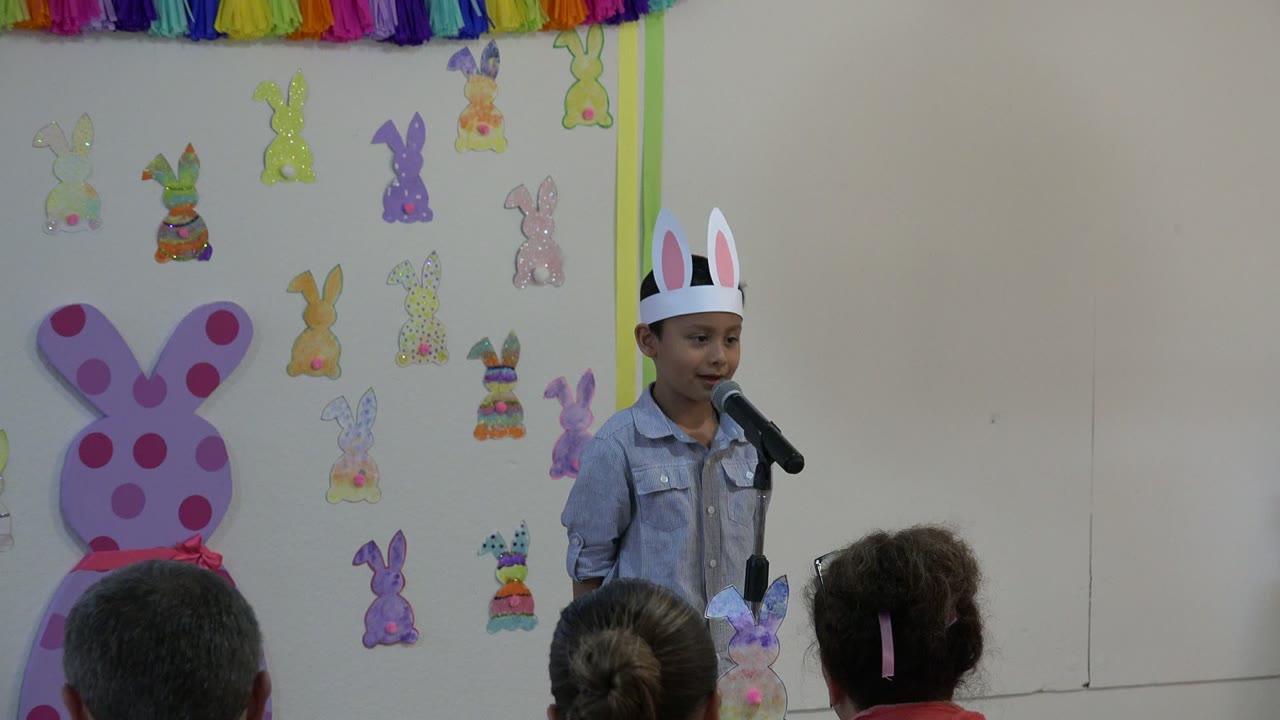 Medium-wide Cabo San Lucas frame of the boy in white bunny ears facing a black microphone as seated adults watch from the foreground beneath paper rabbit decorations.