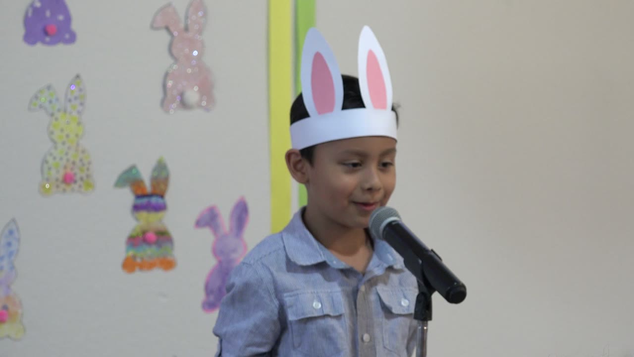A young boy in Cabo San Lucas, Mexico, wears white paper bunny ears and stands beside a black microphone against a wall decorated with pastel bunny cutouts.