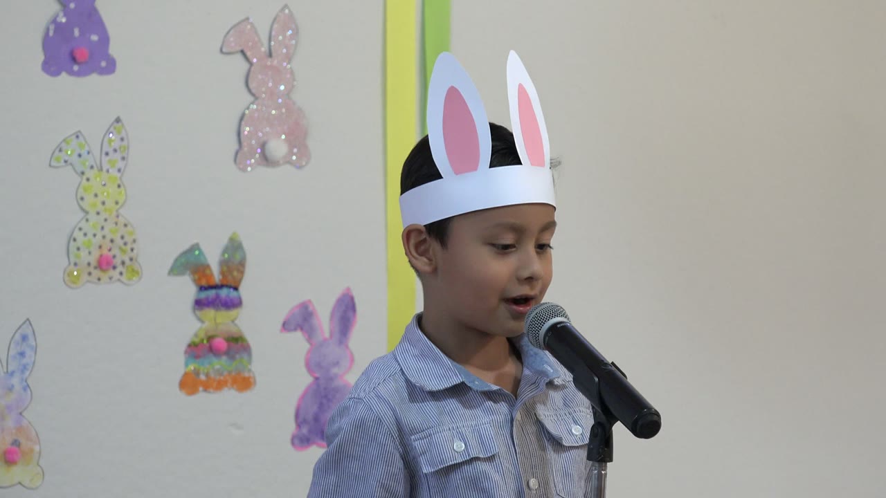 Child wearing paper rabbit ears speaks into a microphone beside rabbit wall cutouts.