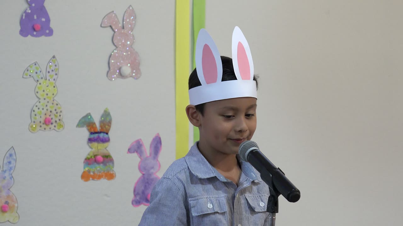 Boy in bunny ears speaking at a microphone with Easter decorations behind him.