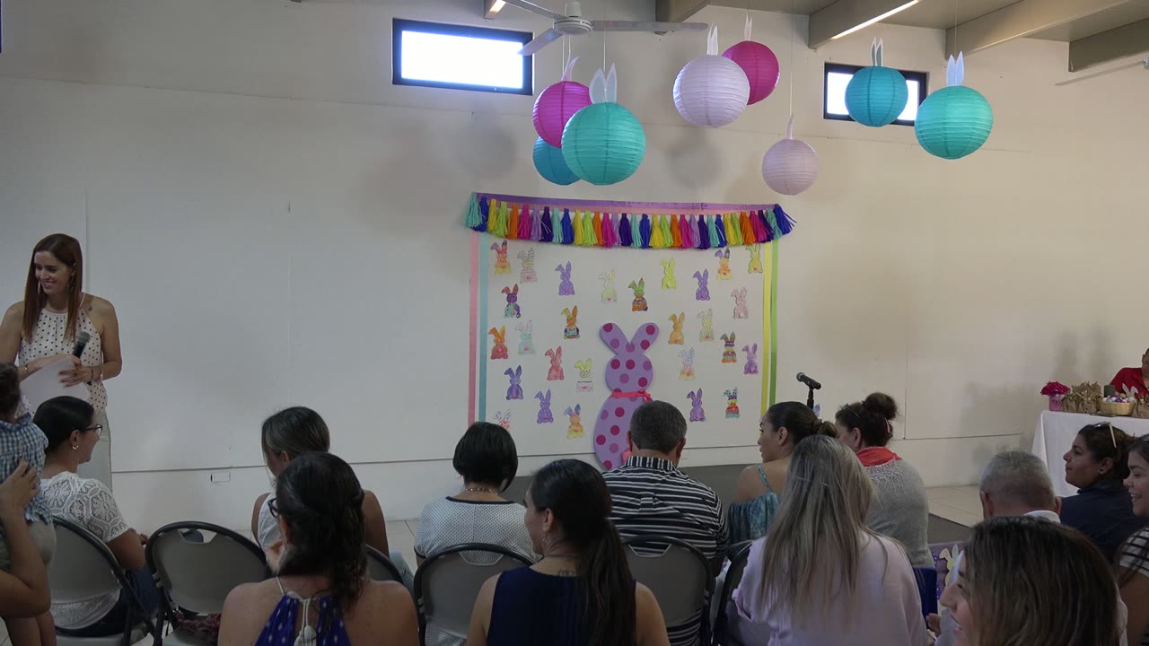 A woman stands at the left edge of an indoor Easter stage in Cabo San Lucas as seated adults and children face a bunny-decorated backdrop.