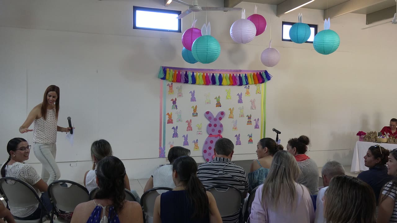 A woman with papers and a handheld microphone stands before the Cabo San Lucas audience, with pastel lanterns and Easter bunny decorations behind her.