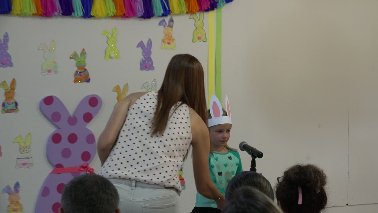 In Cabo San Lucas, Mexico, a young girl wearing white paper bunny ears and a green shirt appears at the microphone while the teacher leans near her in the decorated classroom.