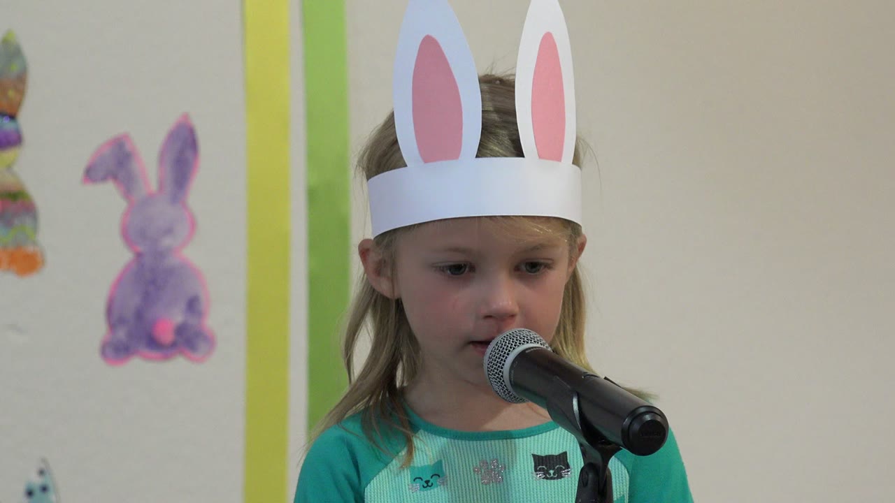 At 0:01 in Cabo San Lucas, Mexico, the girl in white bunny ears looks slightly downward while standing close to the black microphone and decorated wall.