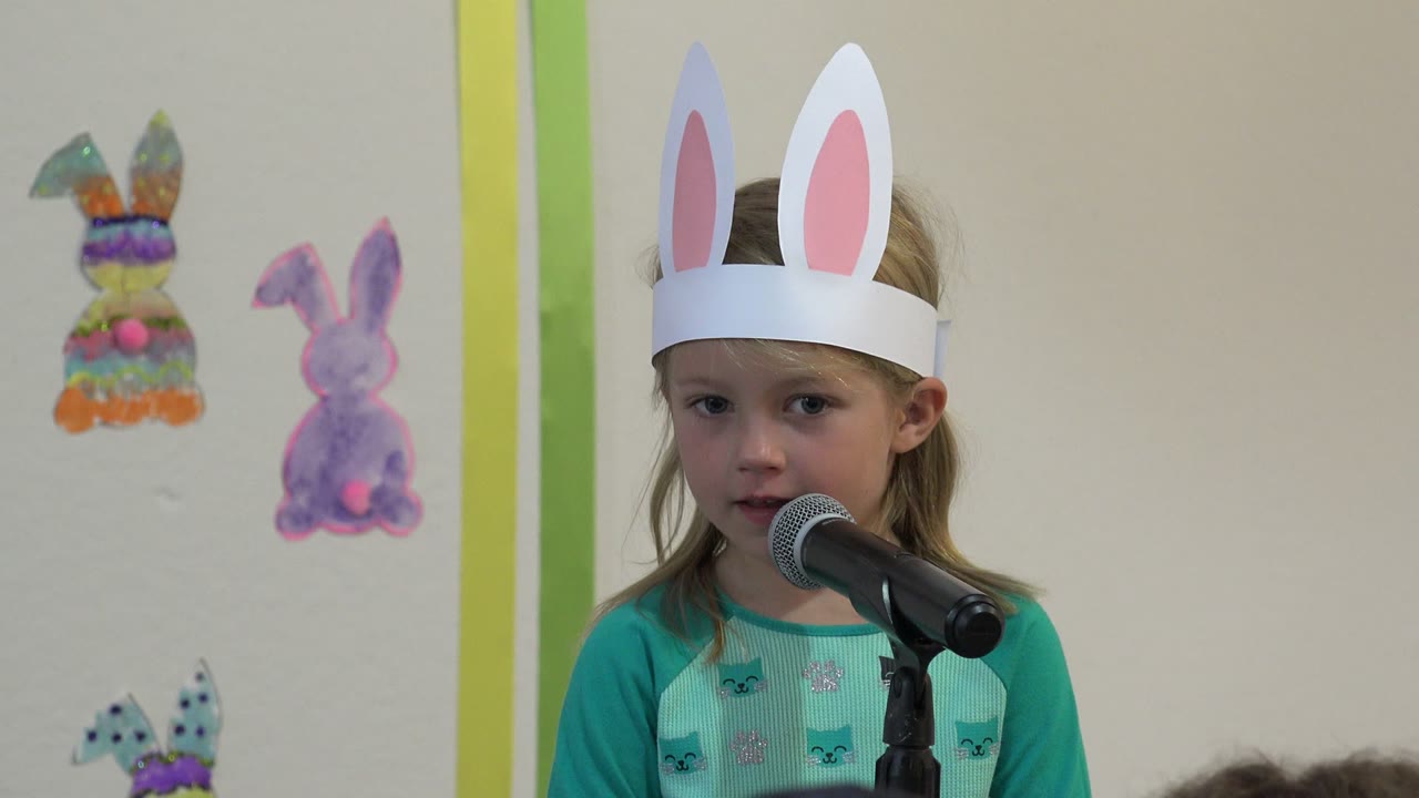 Young girl in white bunny ears faces a microphone during an Easter play in Cabo San Lucas, Mexico, with handmade bunny wall decorations behind her.