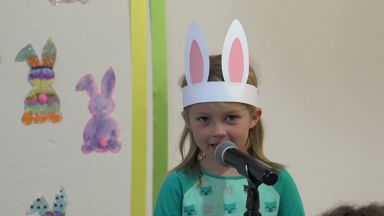 The girl smiles slightly beside the microphone, wearing white paper bunny ears against a wall decorated with pastel rabbit drawings in Cabo San Lucas, Mexico.