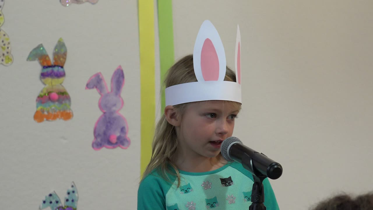 Young girl in white rabbit ears speaking into a black microphone.