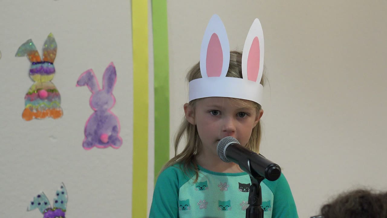 The child looks forward from behind the microphone in Cabo San Lucas, Mexico, with colorful bunny cutouts and vertical green trim in the background.