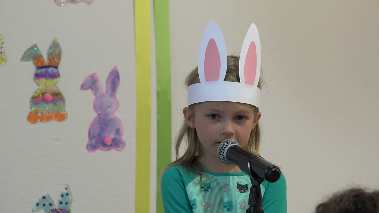 Young girl in paper rabbit ears speaking into a microphone.
