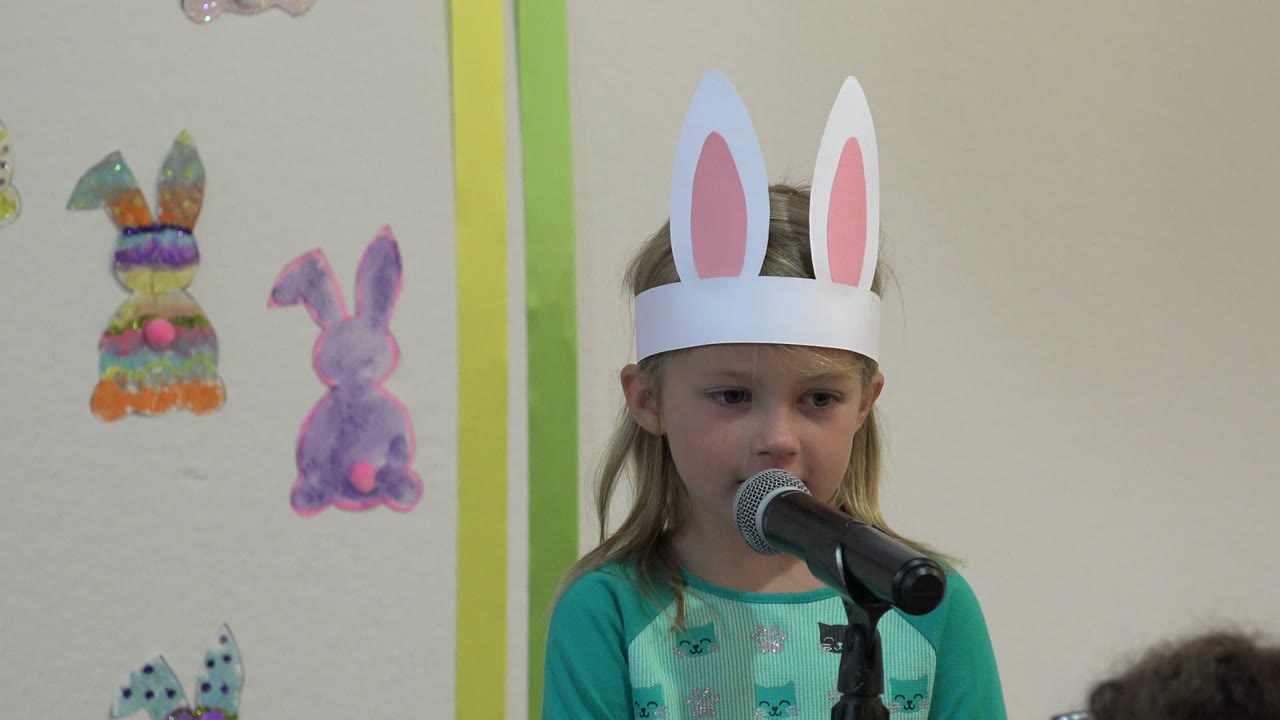 At 0:01, the girl in white rabbit ears looks slightly to the side while standing close to the microphone in the decorated indoor setting in Cabo San Lucas.