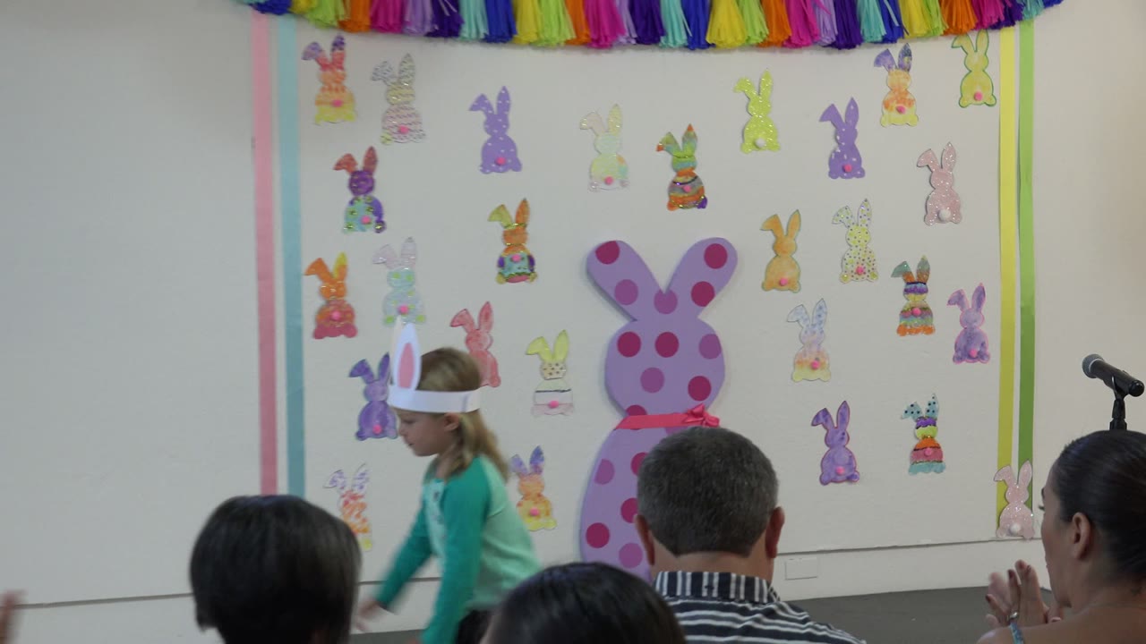 Child in bunny ears moving across an Easter-decorated school stage.