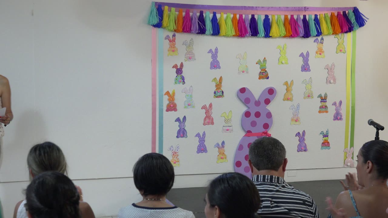 The decorated school stage in Cabo San Lucas, Mexico is centered with paper bunny cutouts, a large pink bunny shape, a microphone stand, and seated audience members in front.