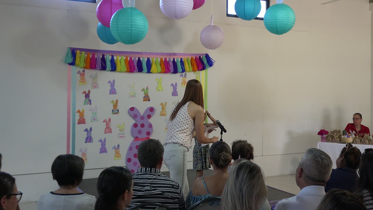 Medium-wide Cabo San Lucas frame with a girl at a microphone, an adult leaning toward her, seated viewers in the foreground, and paper bunny decorations on the wall.