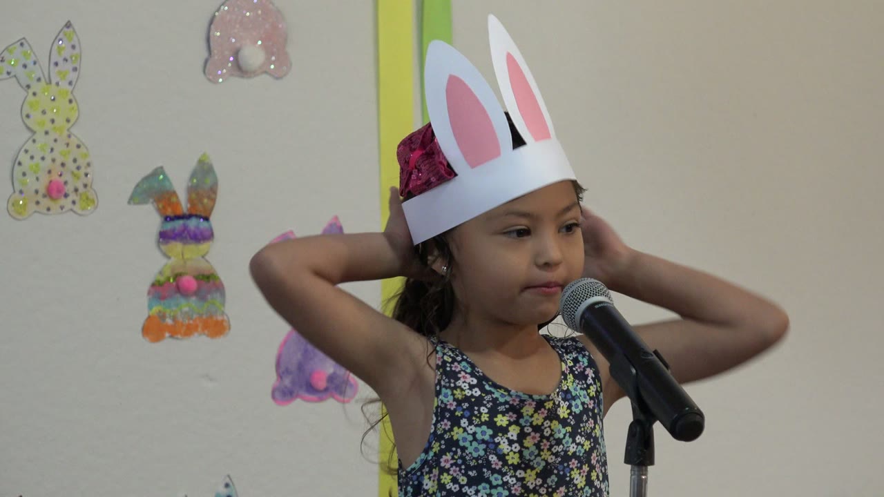Young girl in Cabo San Lucas holds both hands behind her head while standing close to a microphone in front of Easter bunny decorations.