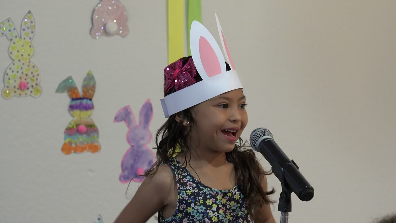 A young girl in Cabo San Lucas smiles beside a black microphone while wearing a white paper bunny-ear crown, with pastel bunny decorations on the wall behind her.