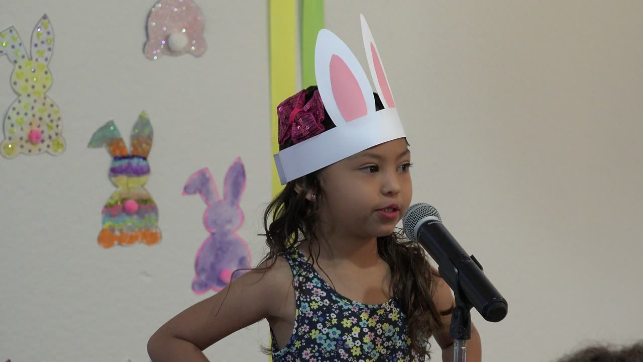 A young girl in Cabo San Lucas faces slightly toward a black microphone while wearing paper bunny ears, with colorful handmade bunny decorations visible on the indoor wall.