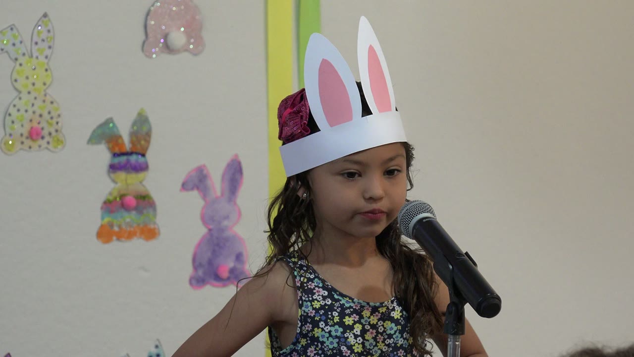 Young girl in bunny-ear crown looking forward by a microphone.
