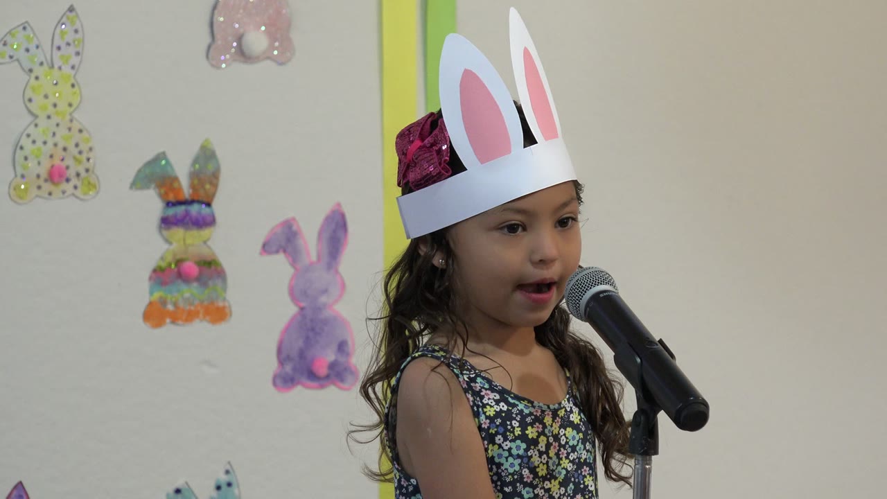 At 0:00 in Cabo San Lucas, Mexico, a young girl wearing paper bunny ears faces a microphone with Easter bunny decorations on the wall behind her.
