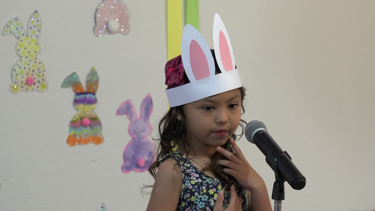 Young girl in paper rabbit ears beside a microphone indoors.
