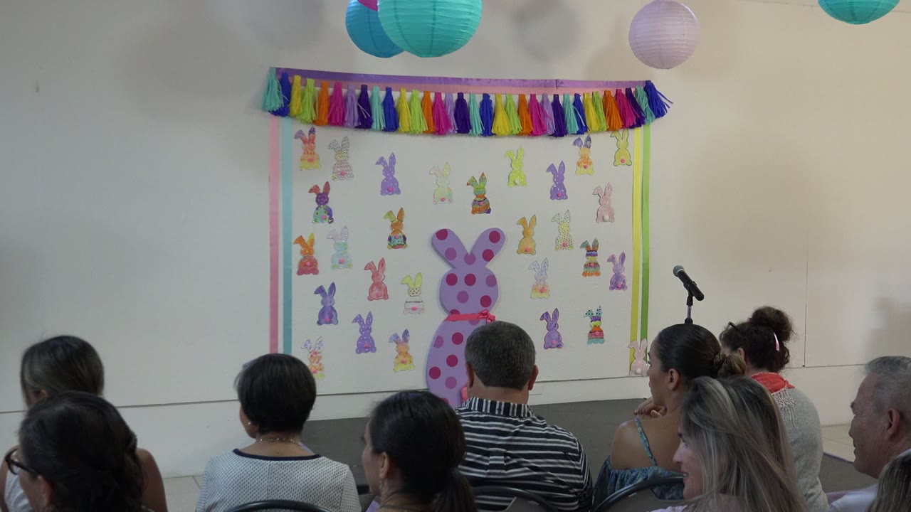 The second frame shows the same Cabo San Lucas school event room with adults in chairs, teal and purple paper lanterns overhead, and a bunny-themed display on the stage wall.
