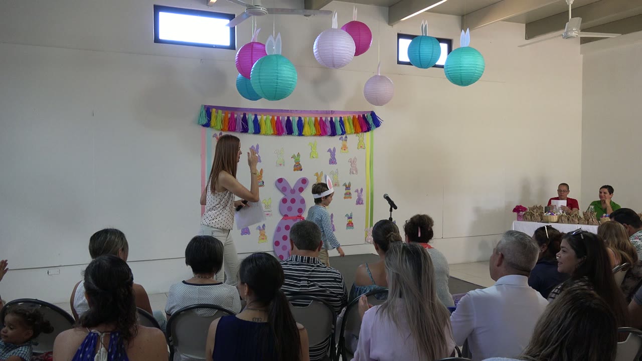The woman stands closer to the decorated wall as the boy in rabbit ears remains beside the black microphone, with audience members seated across the lower frame.