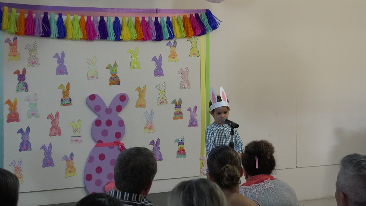 Young child in bunny ears stands at a microphone in front of classroom decorations.