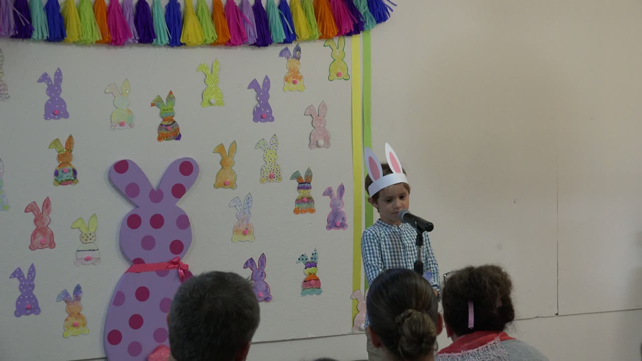 At 0:01, the child remains at the microphone in Cabo San Lucas, Mexico, with the large pink bunny cutout more centered and adults seated below the stage area.