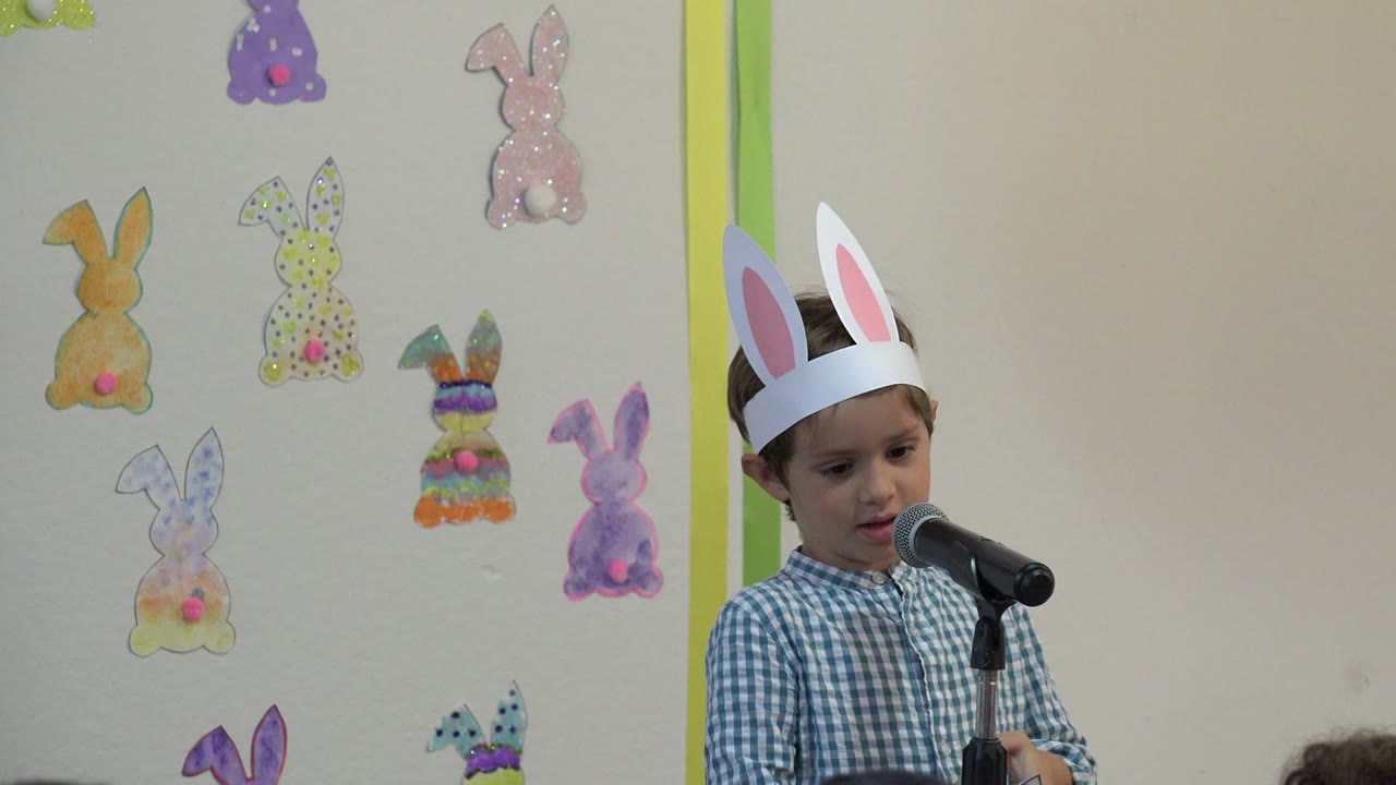 The boy in rabbit ears looks downward near the microphone, with paper bunny cutouts and a green vertical strip on the wall behind him in Cabo San Lucas, Mexico.