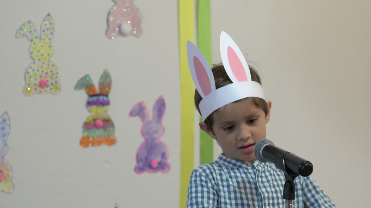 Boy in bunny ears standing at a microphone before Easter decorations.