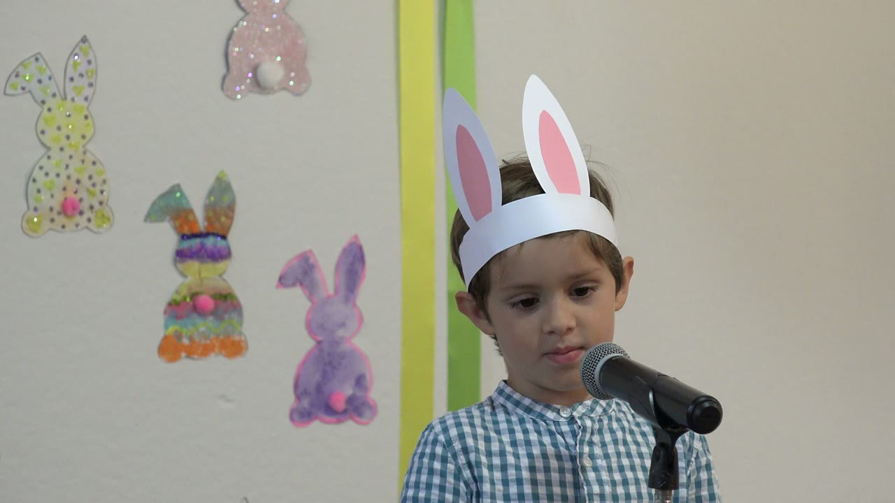 The child in a rabbit-ear headband looks slightly downward while standing by the microphone in front of handmade bunny decorations in Cabo San Lucas.