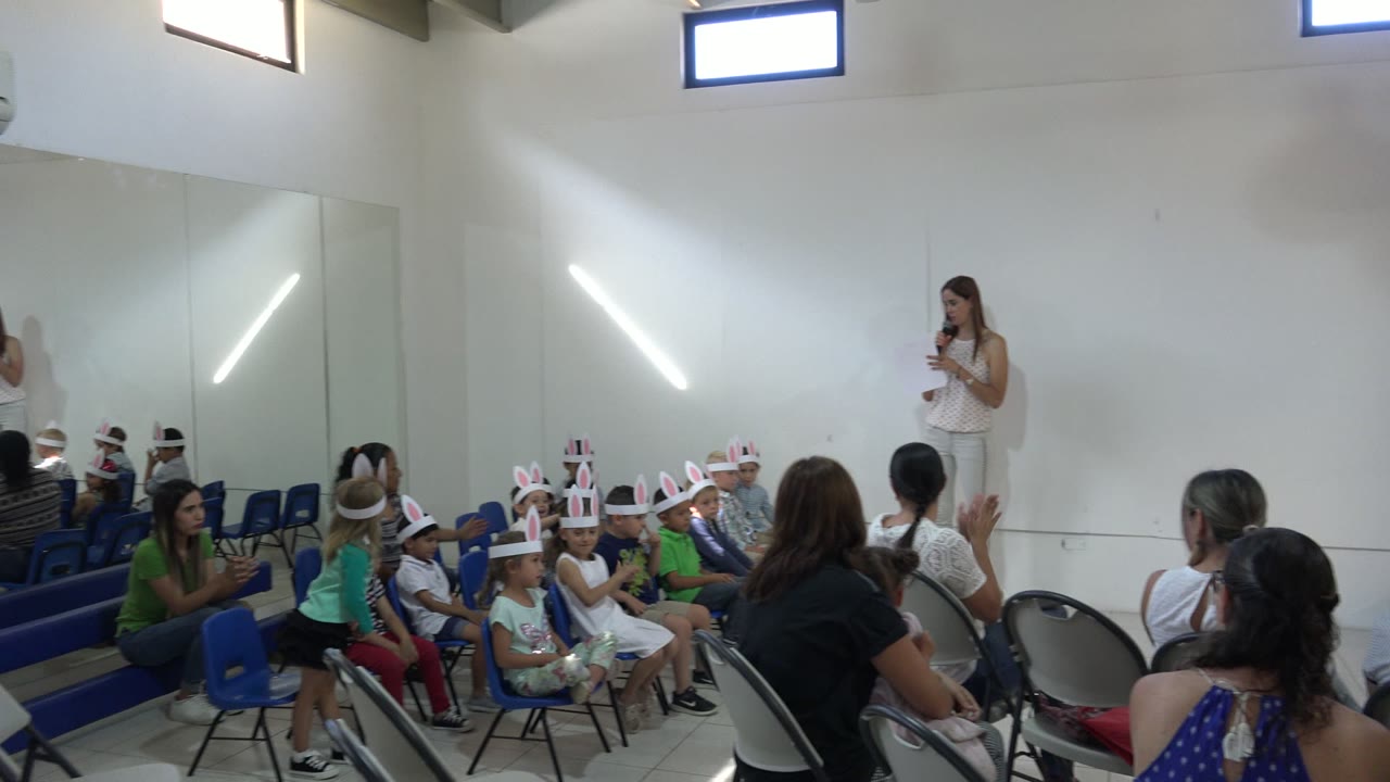 Woman speaking to seated children wearing bunny-ear headbands in an indoor room.