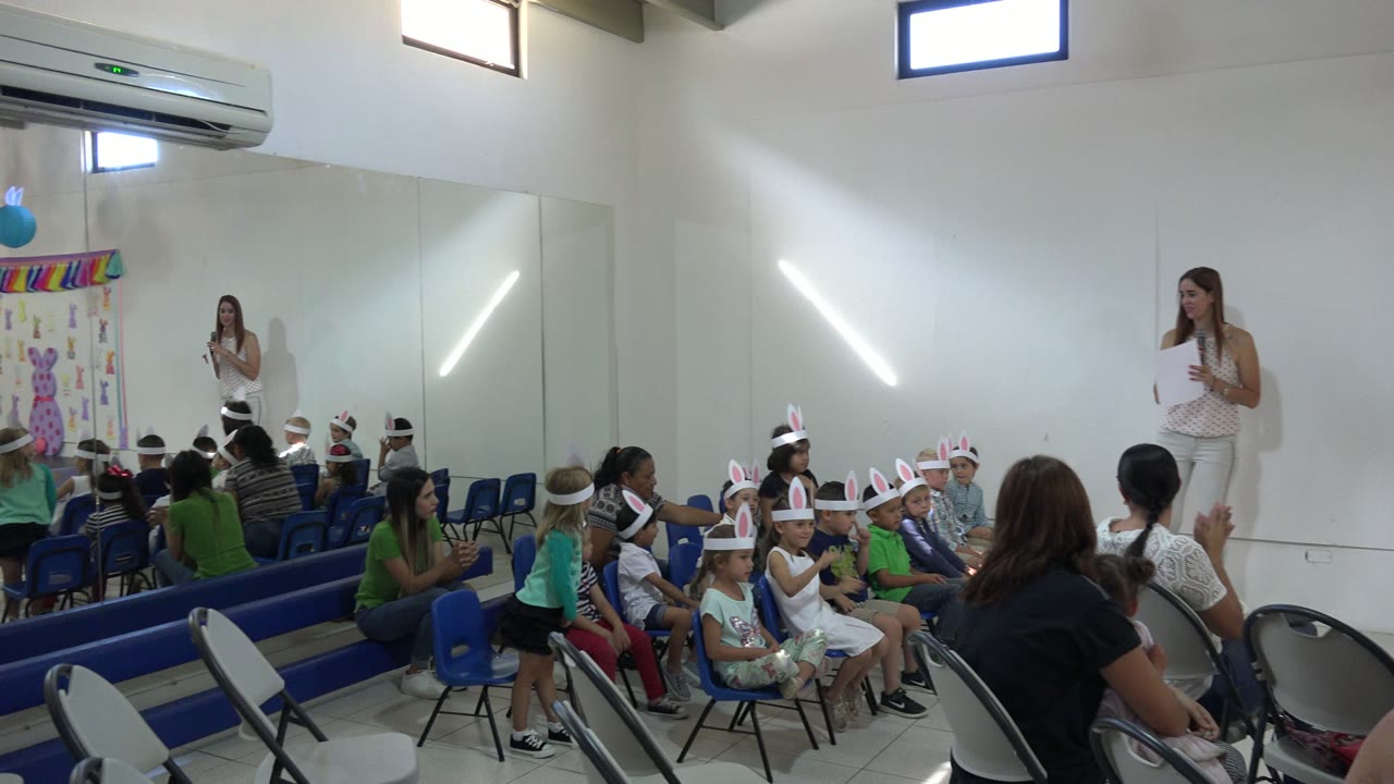 The Cabo San Lucas interior shows the presenter at right, children seated in rows at center, adults nearby, and a decorated backdrop with balloons reflected in the mirror.