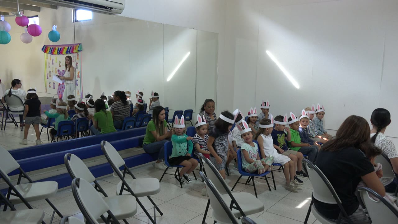 Cabo San Lucas, Mexico frame with children in bunny ear headbands seated in small chairs while adults sit behind them and a decorated Easter backdrop stands at the front of the room.
