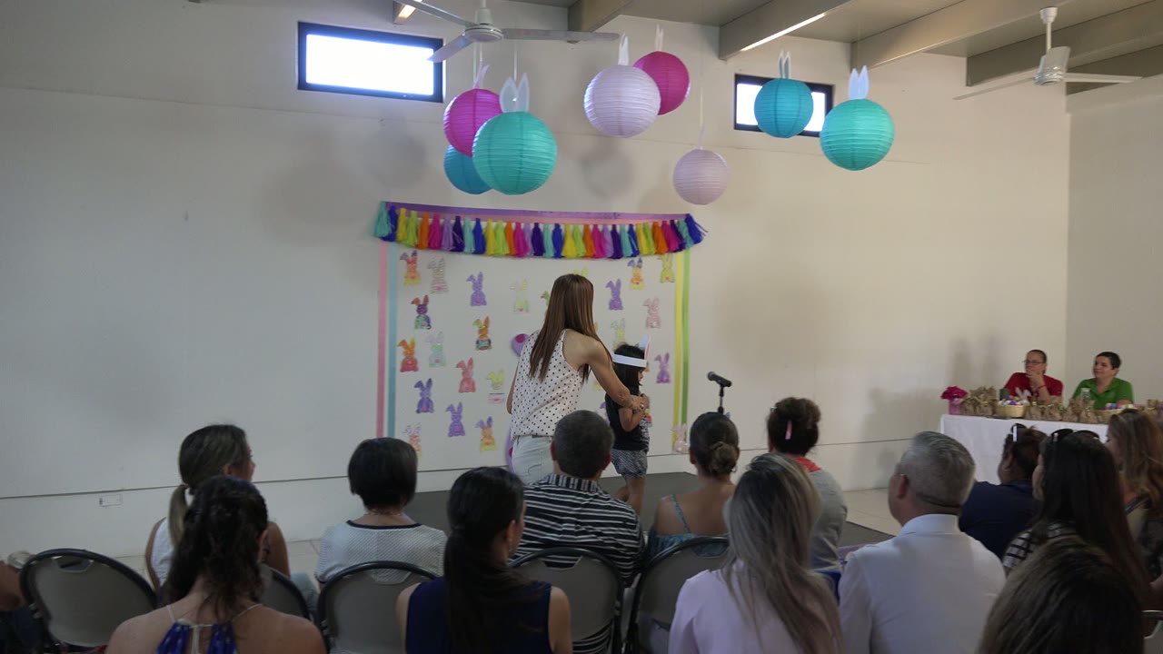 Cabo San Lucas school event frame with a woman standing beside a child at the presentation area while adults sit in rows under colorful hanging lanterns.