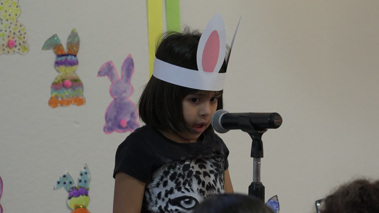 Young girl in Cabo San Lucas wearing paper bunny ears stands close to a black microphone, with colorful bunny decorations on the wall behind her.